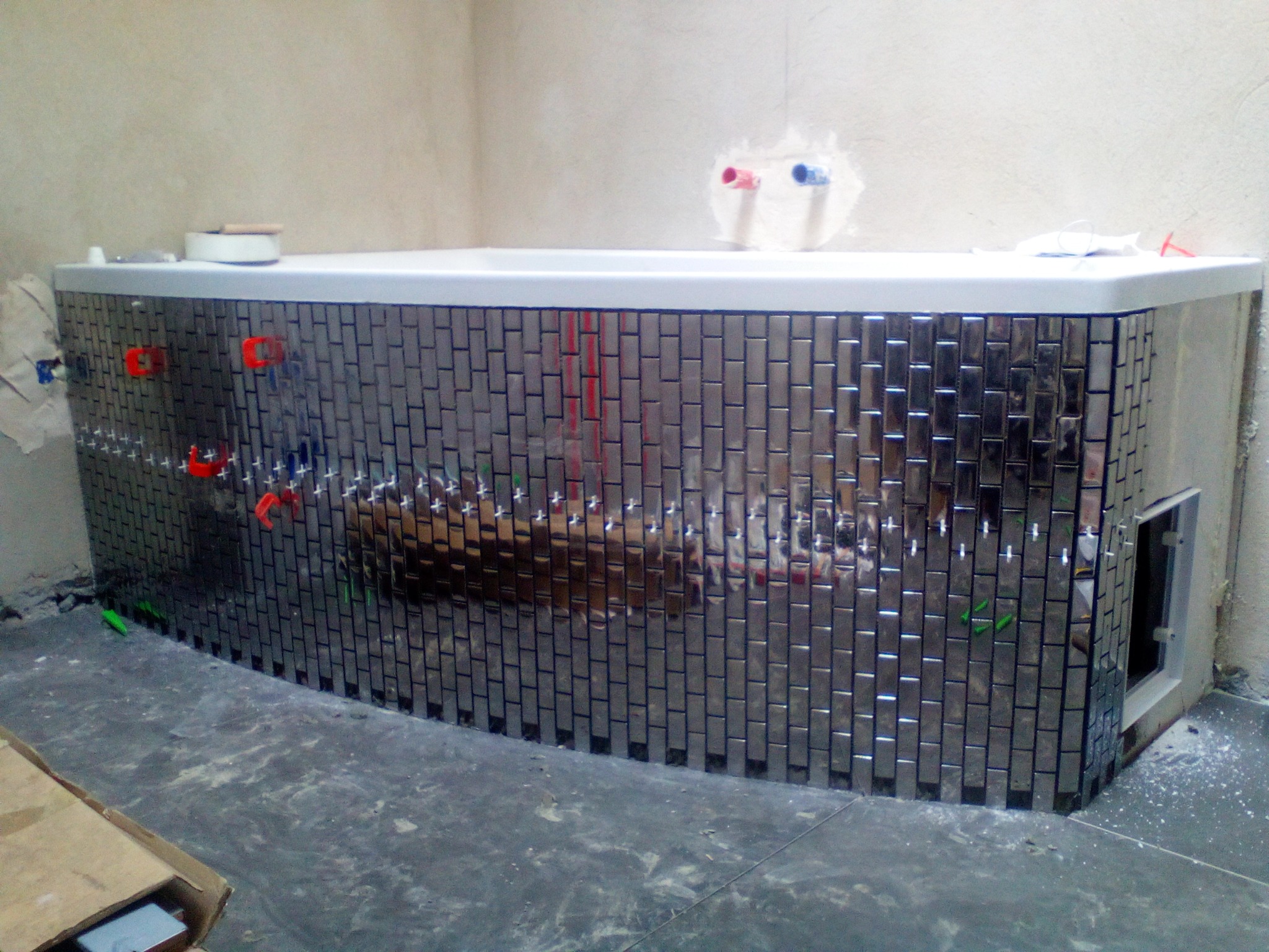 Obudowa wanny wykończona błyszczącą, metaliczną mozaiką w trakcie montażu, widoczne krzyżyki dystansowe i otwór rewizyjny.