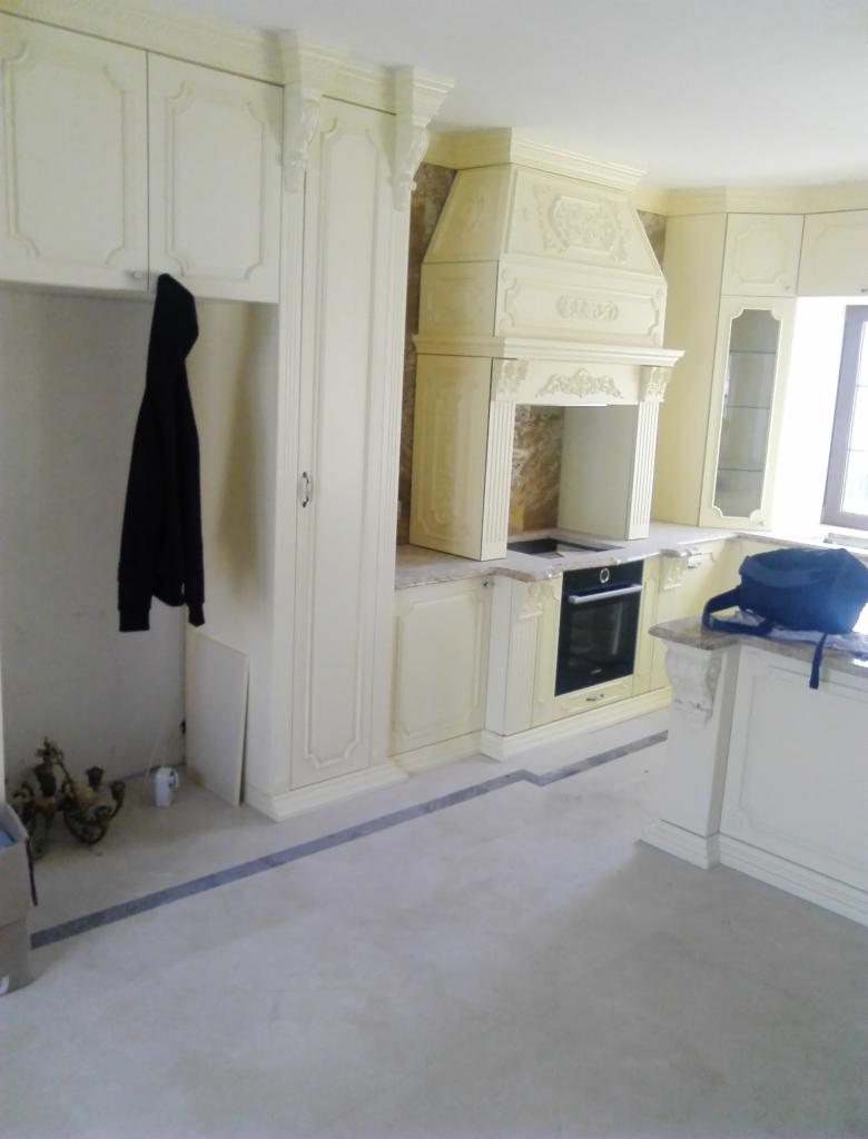 Jasna kuchnia w stylu klasycznym z ozdobnym okapem, wiszącą czarną kurtką i elementami dekoracyjnymi na podłodze.