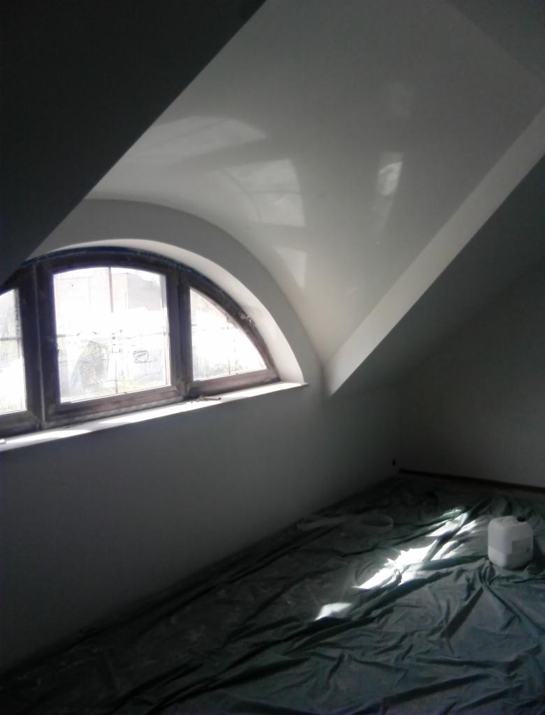 Poddasze ze świeżo pomalowanym sufitem na biało, widoczne okno łukowe i zabezpieczona podłoga folią malarską, kanister z farbą w rogu.