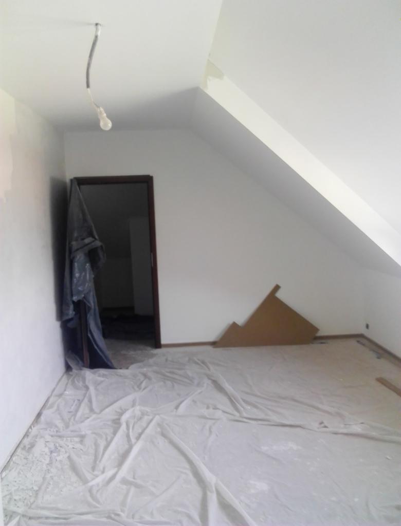 Pokój na poddaszu w trakcie przygotowań do malowania: podłoga zabezpieczona folią malarską, widoczne niedoskonałości na ścianie, wystająca żarówka i fragmenty kartonów przy ścianie.