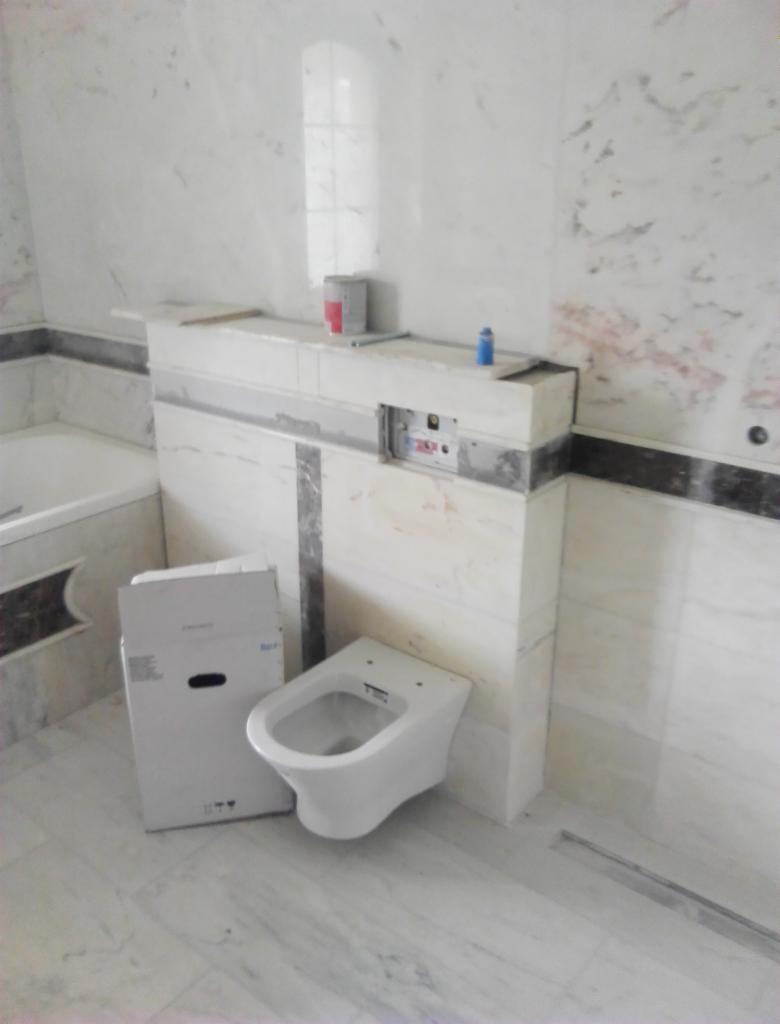 Montaż podwieszanej toalety w łazience wykończonej marmurem, widoczna kartonowa obudowa stelaża, puszka z farbą i niebieska buteleczka na półce.