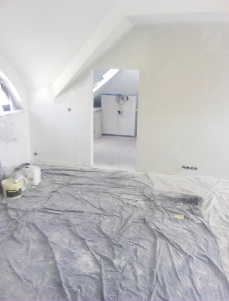 Pokój na poddaszu w trakcie malowania, zabezpieczona folią podłoga, widoczne puszki z farbą i gładzie na ścianach przed ostatecznym wykończeniem.