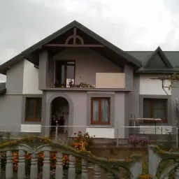 Dwupiętrowy dom z szarą elewacją i ciemnozielonym dachem, widok zza ozdobnego betonowego ogrodzenia porośniętego mchem, na drugim planie rusztowanie przy ścianie.