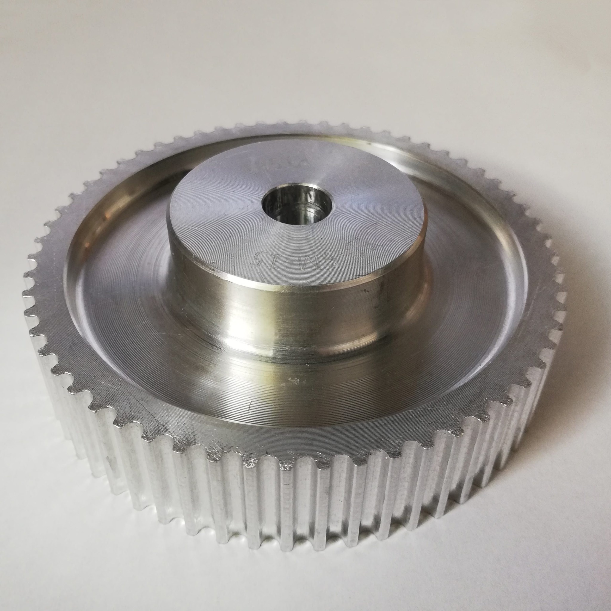 Precyzyjnie wykonane, srebrne koło zębate z wyraźnie widocznymi ząbkami i oznaczeniem 'SM-15' na powierzchni, leżące na jasnym tle.
