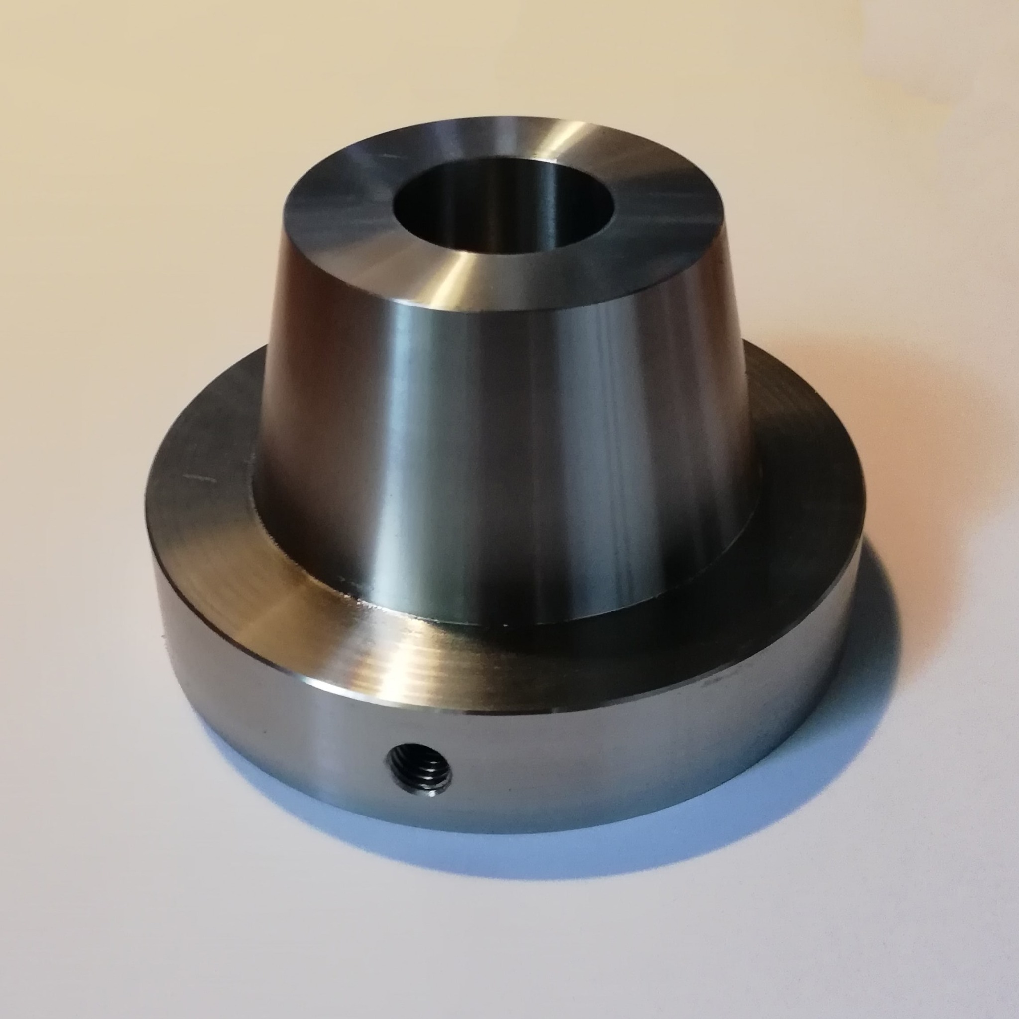 Metalowy element obrobiony na tokarce, z otworem centralnym i gwintowanym bocznym otworem, położony na jasnym tle.