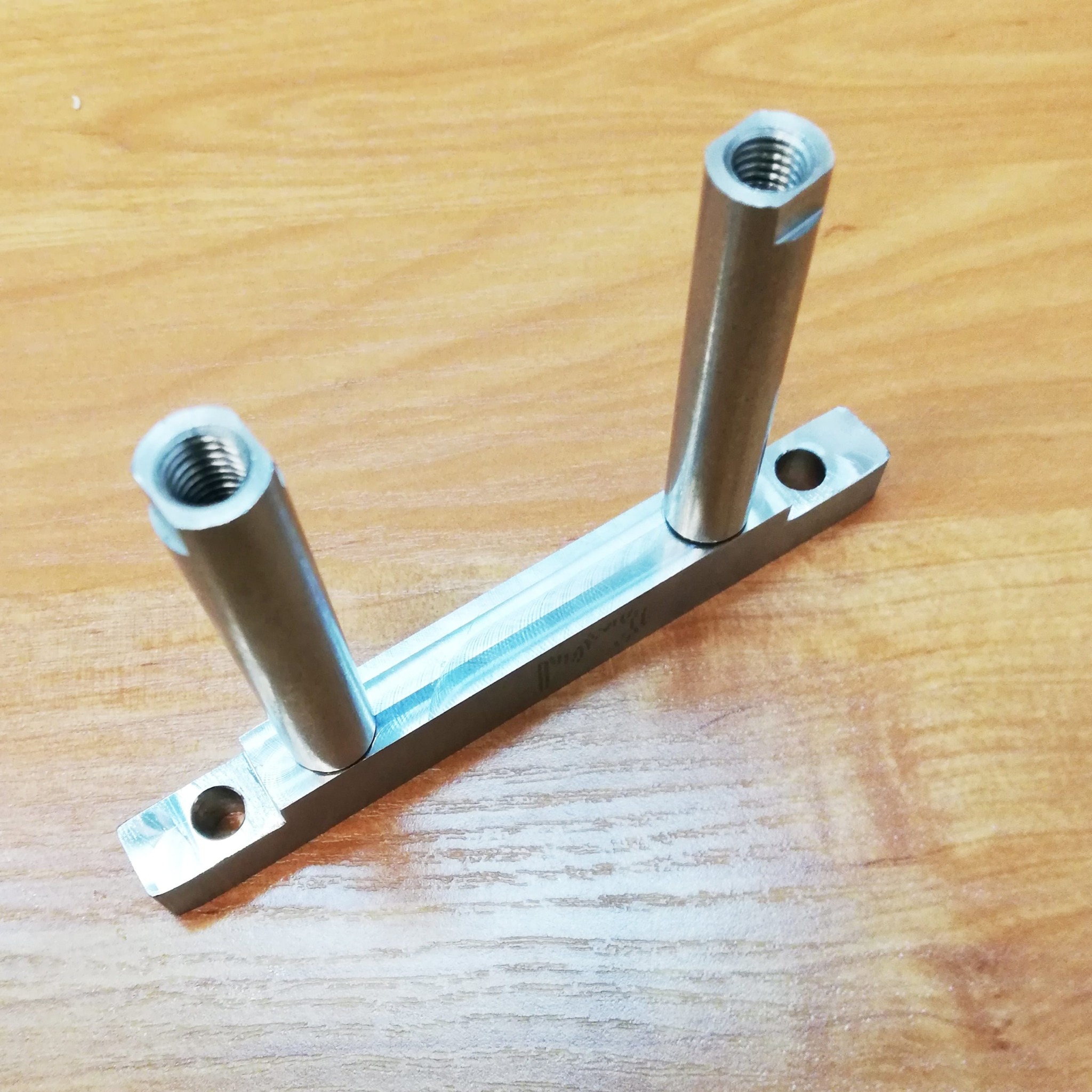Metalowy element konstrukcyjny z dwoma gwintowanymi cylindrami, leżący na drewnianej powierzchni.