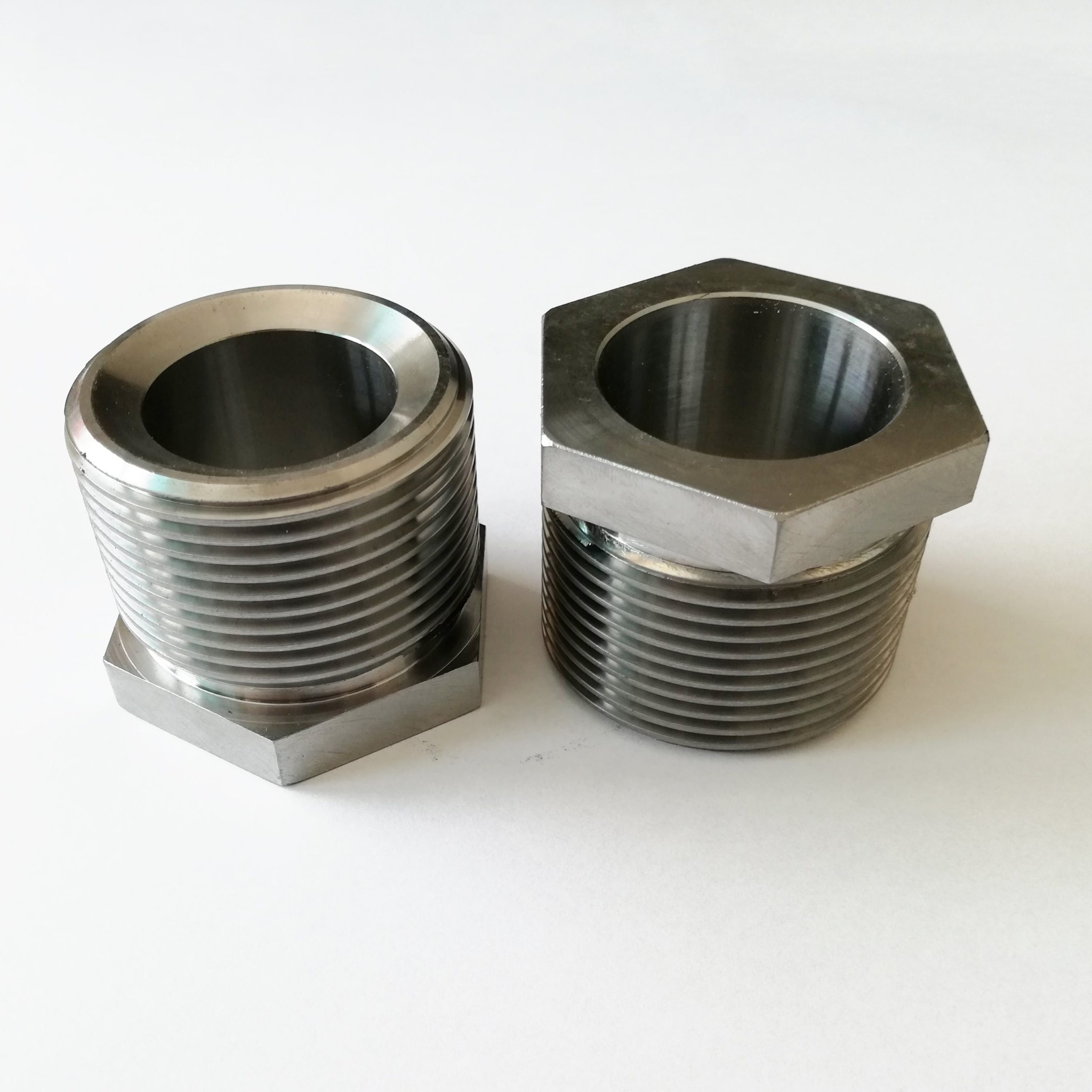 Dwie metalowe tuleje gwintowane o różnych kształtach: okrągła i sześciokątna, leżące obok siebie na białym tle.