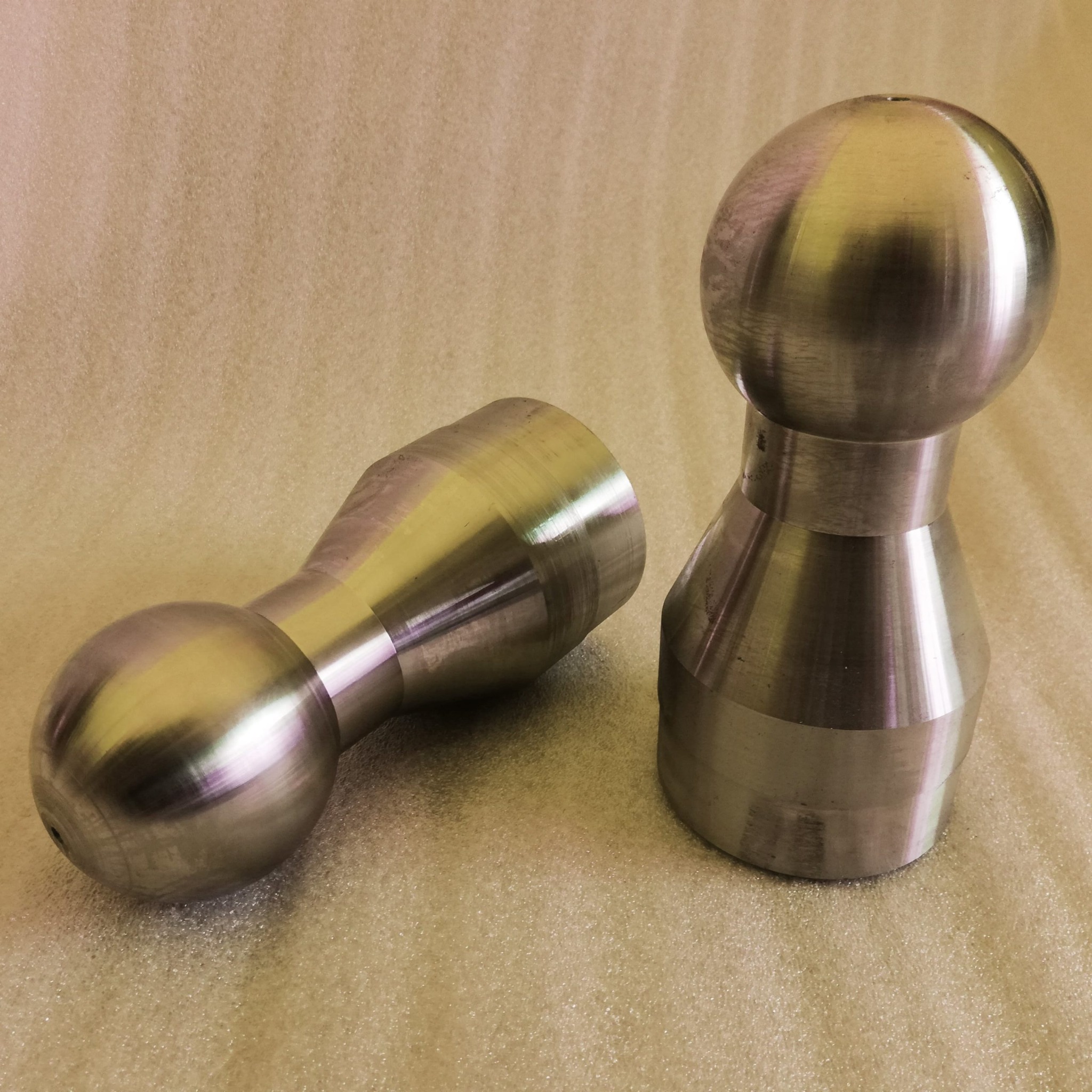 Dwa metalowe elementy obrobione na tokarce, jeden pionowo, drugi leżący, na beżowym tle.