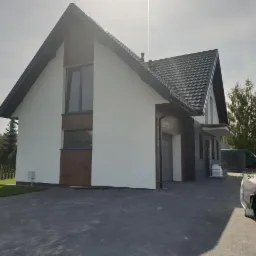 Nowoczesny dom jednorodzinny z białą elewacją i elementami z ciemnego drewna, czarny dach, kostka brukowa na podjeździe, w tle zieleń, widoczne worki i puszki po materiałach budowlanych.