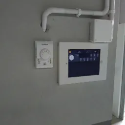 Panel sterowania inteligentnego domu z dotykowym ekranem i regulatorem temperatury Flowair zamontowane na szarej ścianie, widoczne białe rury instalacyjne.