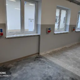 Wnętrze nowo wykończonego pomieszczenia z trzema oknami i zainstalowanymi gniazdami elektrycznymi w kolorach czerwonym i niebieskim, umieszczonymi pod każdym oknem na szarej ścianie.