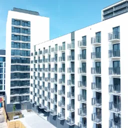 Nowoczesny, biały budynek apartamentowy z symetrycznym układem balkonów, widoczny z góry, z placem budowy i kontenerami w tle.