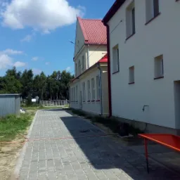 Budynek z czerwoną dach&oacute;wką i chodnikiem z kostki brukowej, widok z poziomu chodnika, błękitne niebo z chmurami w tle.