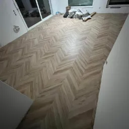 Nowo ułożona podłoga z paneli w jodełkę, widoczne resztki materiałów budowlanych i narzędzia przy oknie, częściowo zasłoniętym żaluzjami.