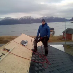 Dekarz w niebieskiej kurtce stoi na połowie dachu pokrytej czarnym gontem bitumicznym, druga połowa dachu pokryta płytami OSB, w tle jezioro i góry ze śniegiem, widać narzędzia dekarskie.