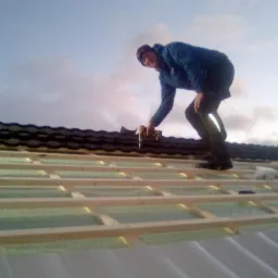 Pracownik w kombinezonie mocuje elementy konstrukcji dachu do drewnianego szkieletu za pomocą wkrętarki na tle pochmurnego nieba.