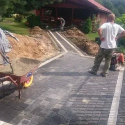 Widok z perspektywy pieszego na nowo ułożoną ścieżkę z ciemnoszarej kostki brukowej z jasnymi obrzeżami, prowadzącą do drewnianego domku, z pracującym mężczyzną w tle i taczką z piaskiem po lewej...