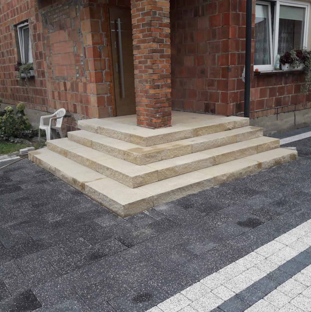 Granitowe schody wejściowe do domu z cegły, otoczone brukowaną nawierzchnią w odcieniach szarości i bieli.