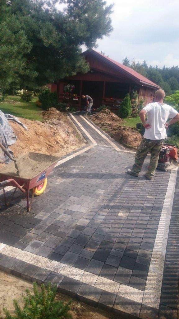 Widok z perspektywy pieszego na nowo ułożoną ścieżkę z ciemnoszarej kostki brukowej z jasnymi obrzeżami, prowadzącą do drewnianego domku, z pracującym mężczyzną w tle i taczką z piaskiem po lewej...