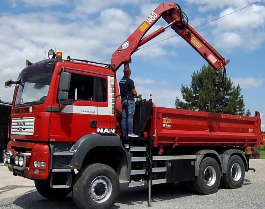 Czerwona ciężarówka MAN z dźwigiem hydraulicznym w trakcie pracy, operator stoi na podeście, błękitne niebo w tle.