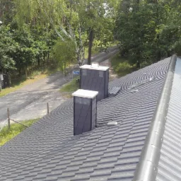 Nowy, grafitowy dach z dachówki ceramicznej z dwoma kominami w kolorze antracytu, widok z góry na tle lasu i drogi.