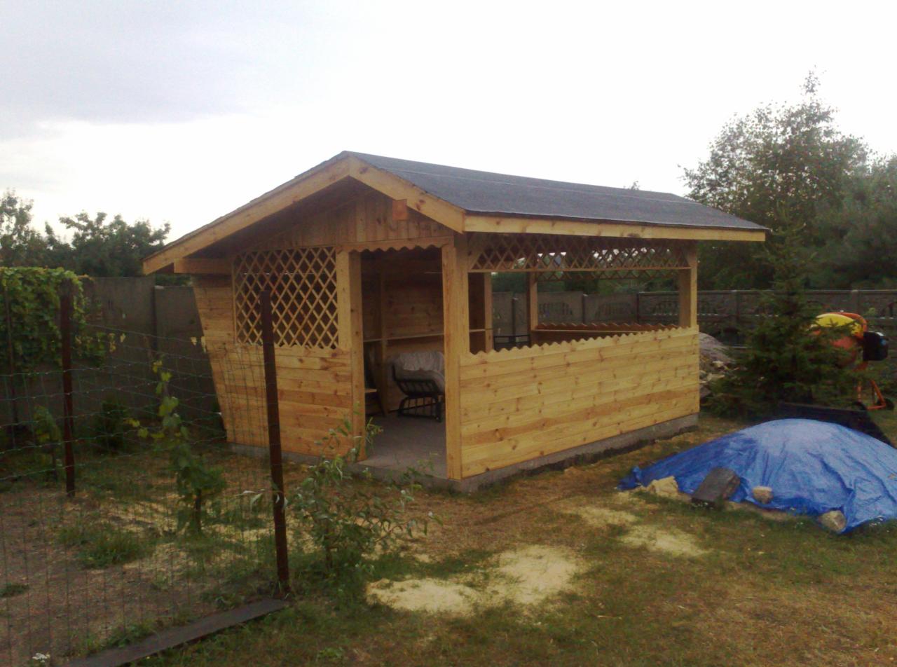 Drewniana altana ogrodowa z czarnym dachem, wykończona kratownicą, z widoczną wewnątrz ławką, na trawiastym terenie, z betoniarką i plandeką w tle.