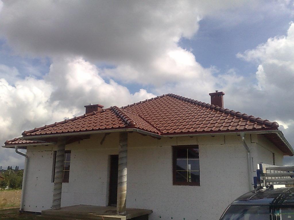 Dom jednorodzinny w trakcie ocieplania z nowym, czerwonym dachem z dachówki ceramicznej i zamontowanymi rynnami, widok na elewację i fragment samochodu.
