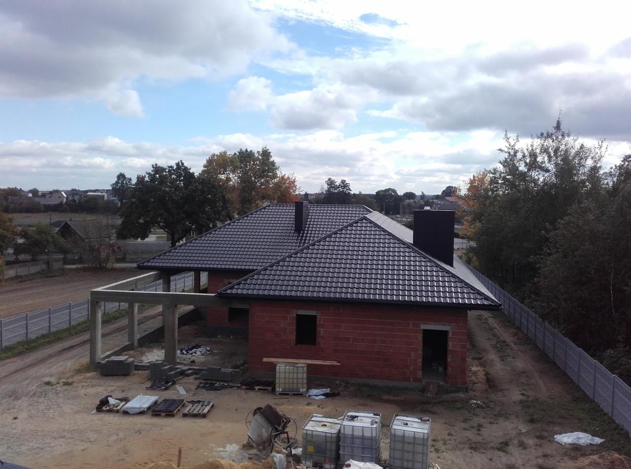 Dom w trakcie budowy z ciemnoszarą dachówką, widoczne materiały budowlane, betonowe słupy werandy i ceglana konstrukcja budynku.