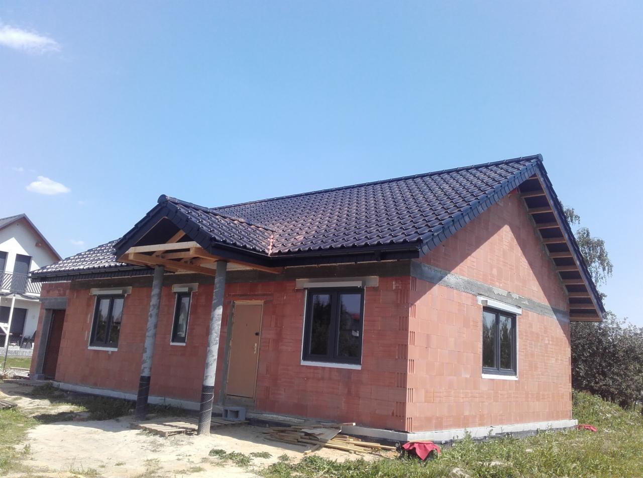 Dom w trakcie budowy z nowym, ciemnym dachem z dachówki ceramicznej i drewnianą konstrukcją zadaszenia wejścia, widoczne rynny i surowy stan elewacji z czerwonej cegły.