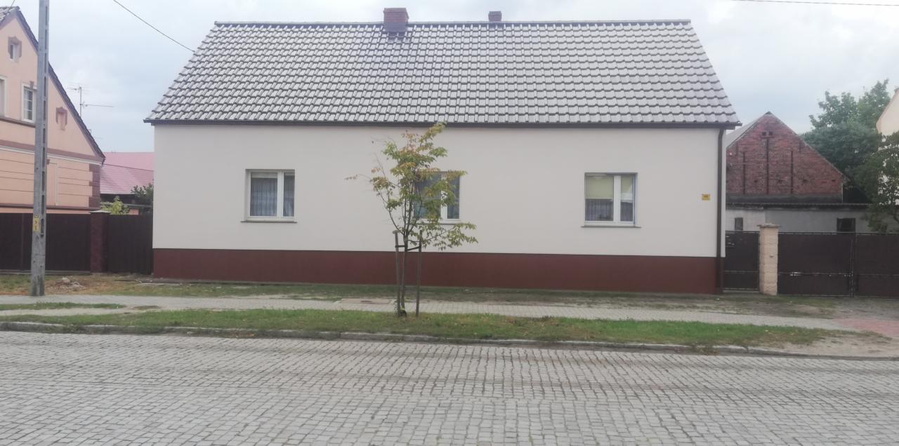 Ocieplony dom jednorodzinny z szarą dachówką, brązowym cokołem i młodym drzewkiem przed elewacją.