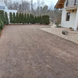 Przygotowany teren pod trawnik z rolki. W tle rząd tuj i budynek. Na pierwszym planie wał do trawy, narzędzia ogrodnicze i fragment kostki brukowej.