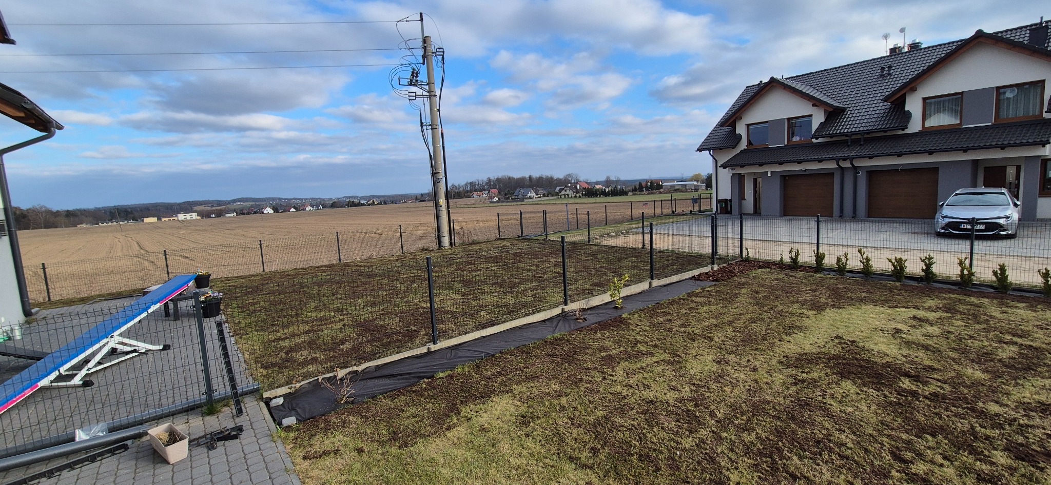Świeżo założony trawnik z młodymi drzewkami wzdłuż ogrodzenia, na tle domu z dwoma garażami i rozległego pola uprawnego. Widoczny rozsypany nawóz.