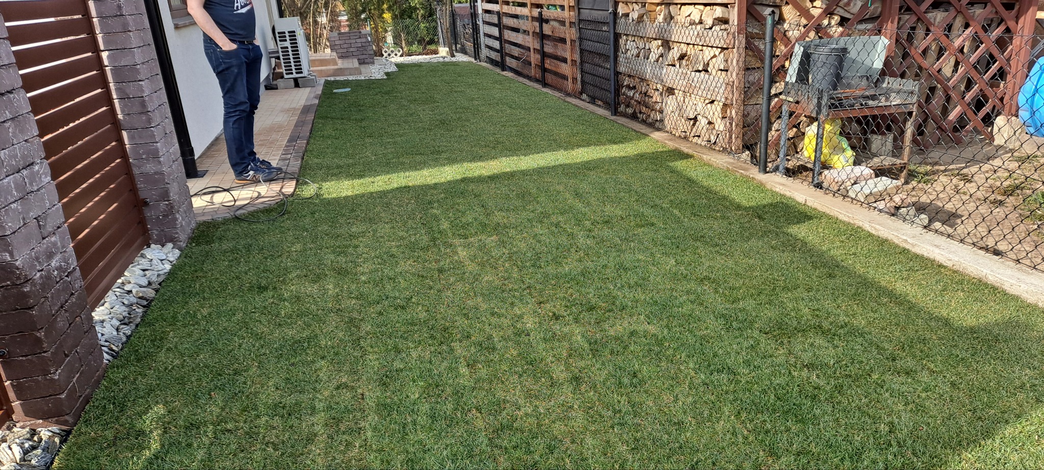 Perfekcyjnie ułożona trawa z rolki w ogrodzie, widoczna część osoby, drewniane elementy architektury ogrodowej w tle, równa powierzchnia trawnika.