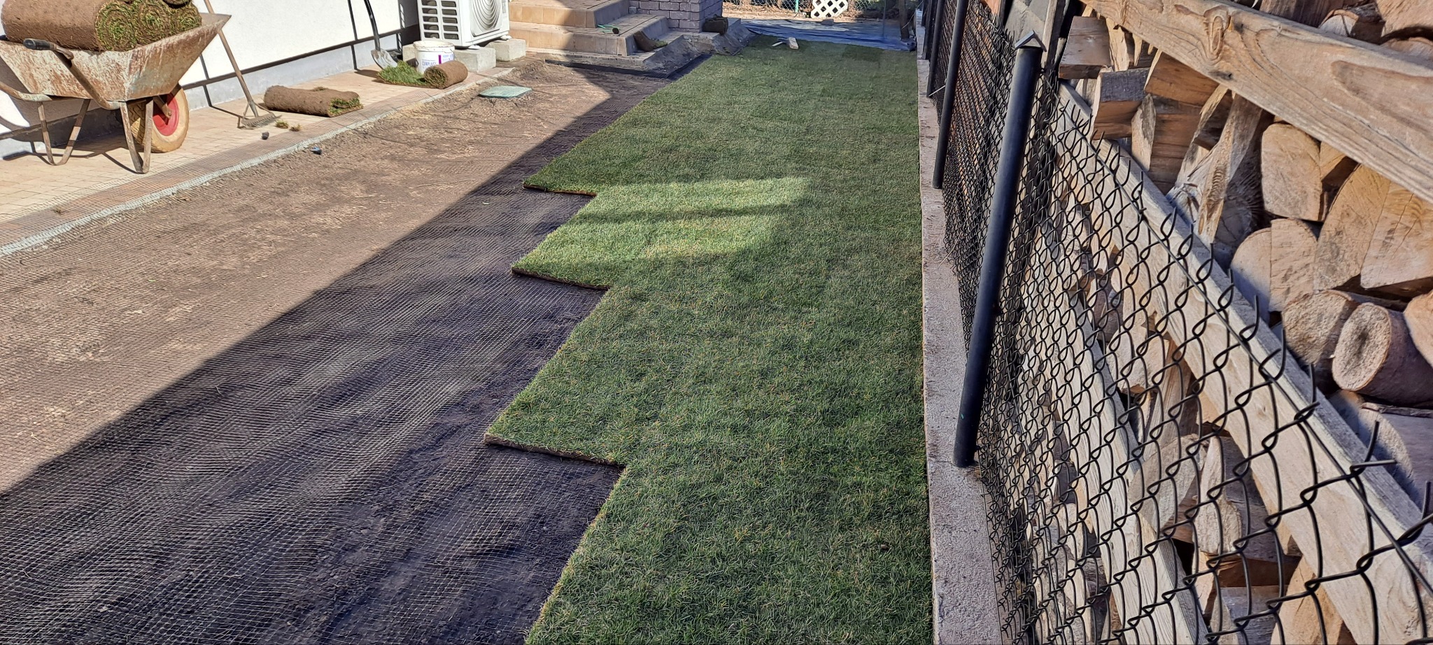 Układanie trawy z rolki: widok z góry na fragment ogrodu z rozłożoną trawą, z rolkami trawy w tle i siatką pod spodem. Widoczny wózek z trawą.