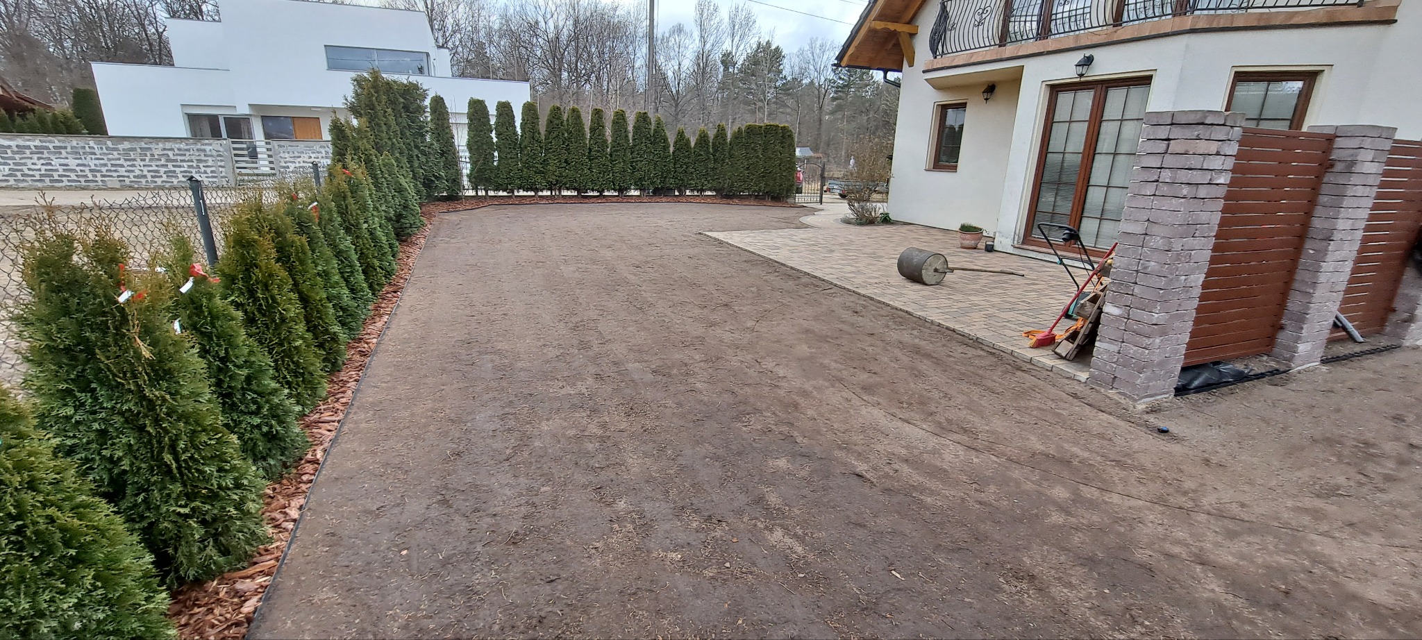 Przygotowany teren pod trawnik z rolki. W tle rząd tuj i budynek. Na pierwszym planie wał do trawy, narzędzia ogrodnicze i fragment kostki brukowej.