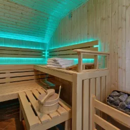 Jasne drewniane wnętrze sauny z oświetleniem LED, wiaderkiem z chochlą, stosem ręczników i piecem z kamieniami.