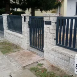 Nowoczesne ogrodzenie z metalowymi przęsłami i słupkami z jasnego kamienia, furtka zamykana na zamek, skrzynka pocztowa z numerem 8AB, widok z ulicy, drzewo w tle.