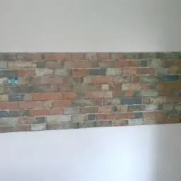 Fragment ściany z tapetą imitującą mur ceglany, z widocznym gniazdkiem elektrycznym, górna i dolna część ściany pomalowana na biało.