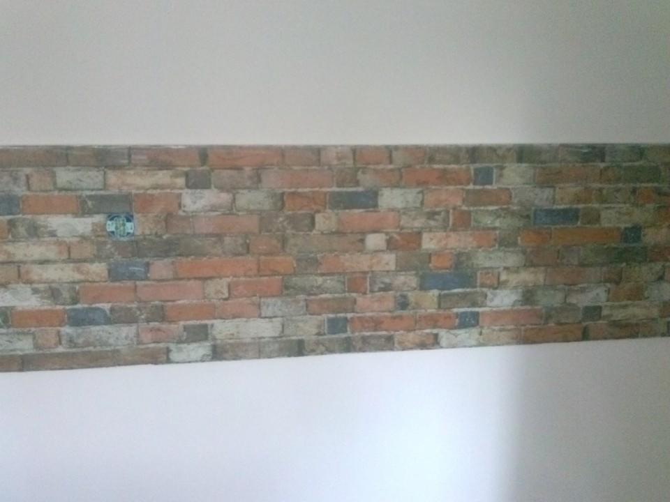 Fragment ściany z tapetą imitującą mur ceglany, z widocznym gniazdkiem elektrycznym, górna i dolna część ściany pomalowana na biało.