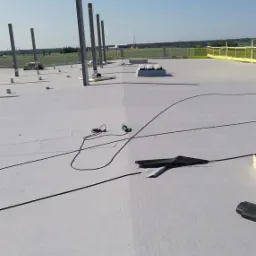 Biała, płaska powierzchnia dachu z widocznymi pionowymi, metalowymi słupami konstrukcyjnymi, przewodami i narzędziami, na tle błękitnego nieba i zieleni.