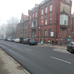 Przedsiębiorstwo Wielobranżowe MEGANT - Ceglany budynek w stylu historyzmu, z widocznymi oknami, balkonem i elementami dekoracyjnymi. Ulica z zaparkowanymi autami i przechodniami w tle, mglista pogoda.