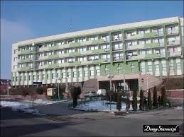 Wielopiętrowy budynek z oknami PCV, jasną elewacją i zielonymi akcentami, sfotografowany w słoneczny dzień z lekko ośnieżonym terenem.