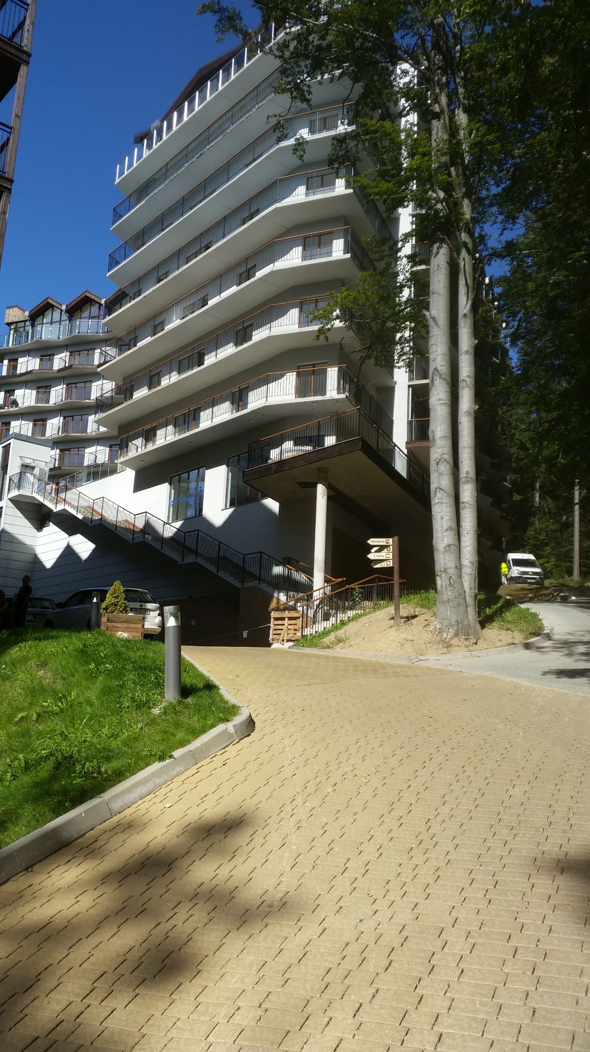 Nowoczesny budynek apartamentowy z balkonami, widok z poziomu brukowanej ścieżki, zieleń i drzewa w otoczeniu. Jasne, słoneczne oświetlenie.