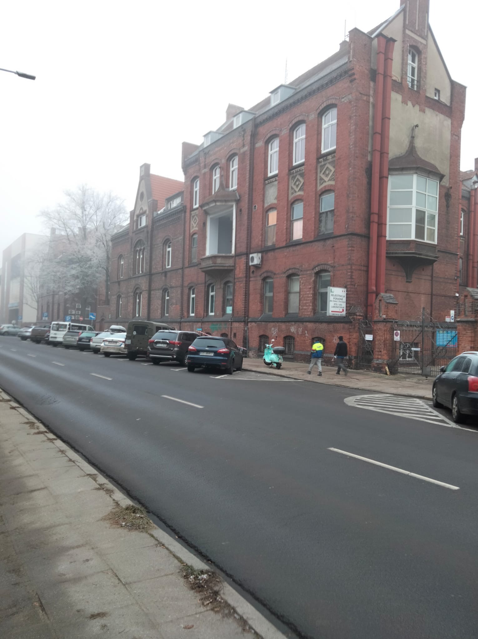 Ceglany budynek w stylu historyzmu, z widocznymi oknami, balkonem i elementami dekoracyjnymi. Ulica z zaparkowanymi autami i przechodniami w tle, mglista pogoda.