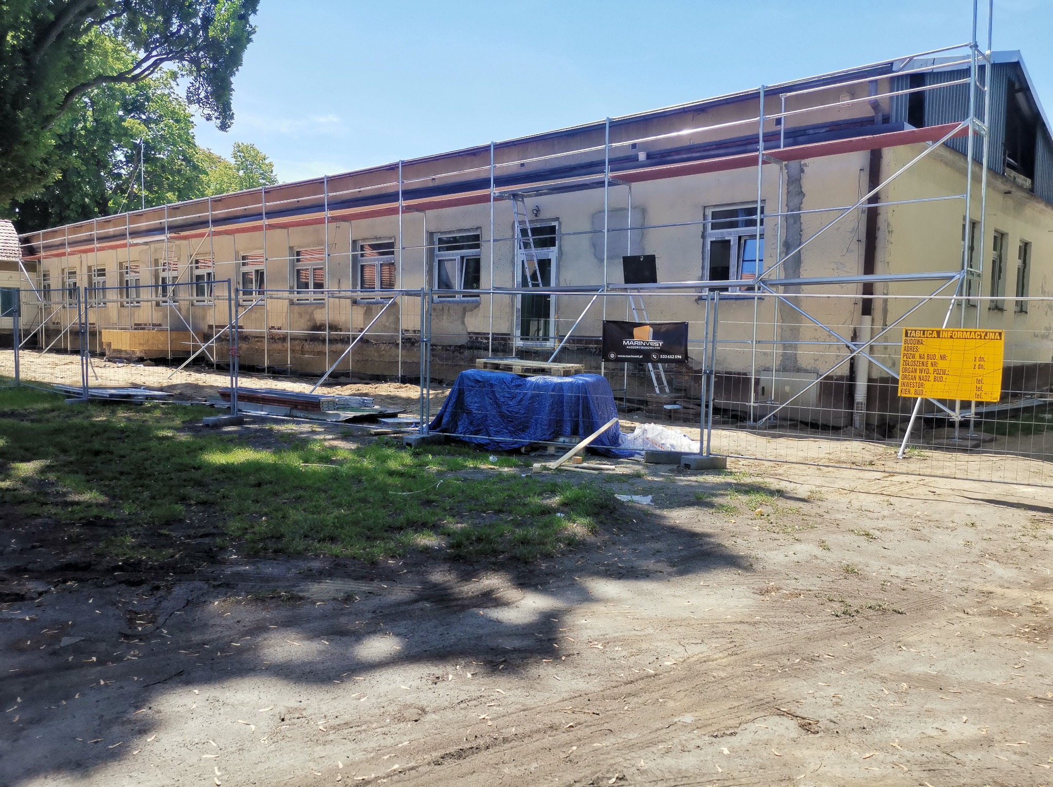 Budynek w trakcie renowacji elewacji, otoczony rusztowaniami i zabezpieczony siatką. Widoczna tablica informacyjna budowy. Prace w toku, teren częściowo zagospodarowany.
