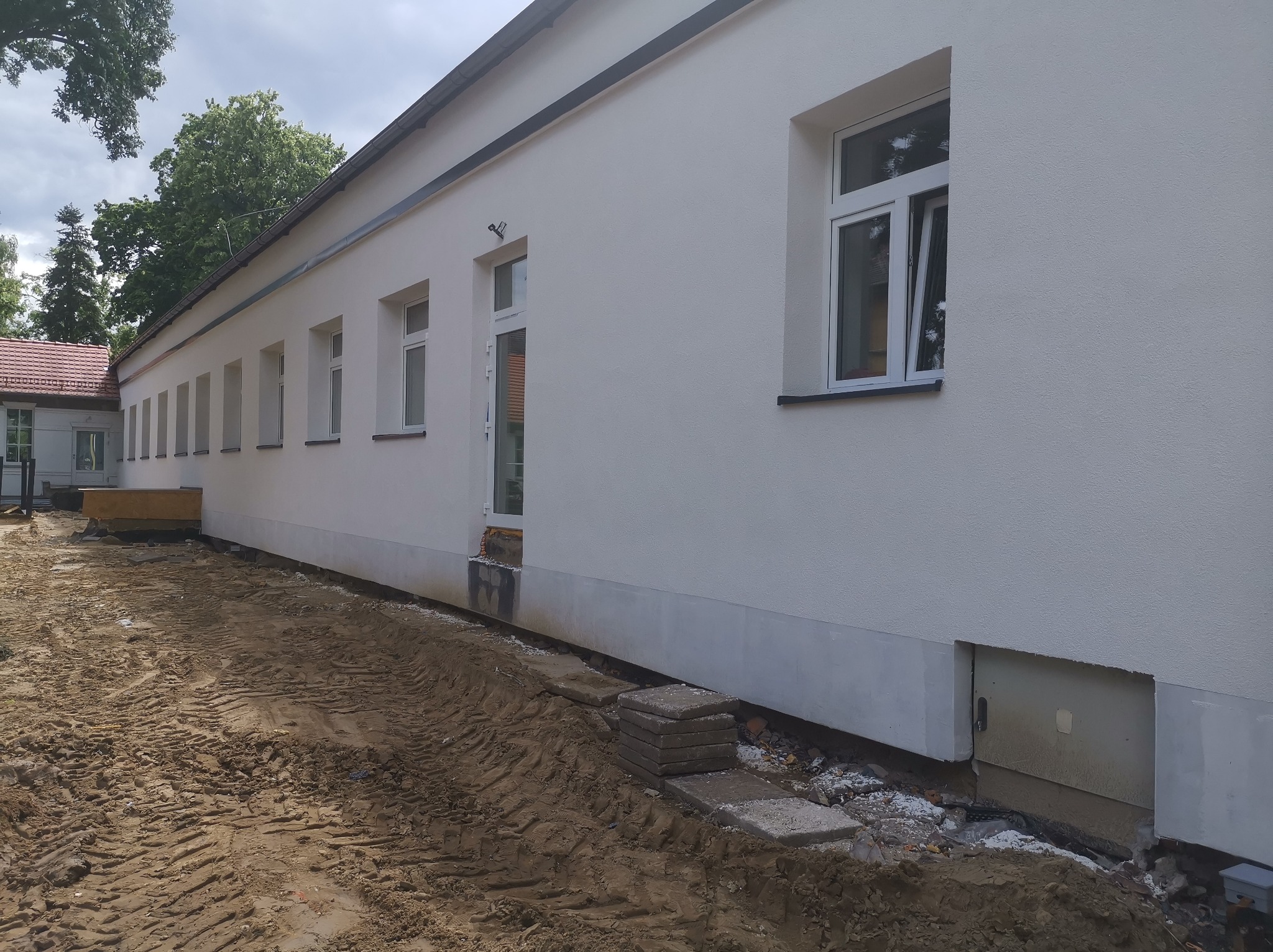 Biała elewacja budynku z nowymi oknami i drzwiami. Widoczne ślady prac ziemnych i przygotowania do dalszych robót wykończeniowych wokół budynku.