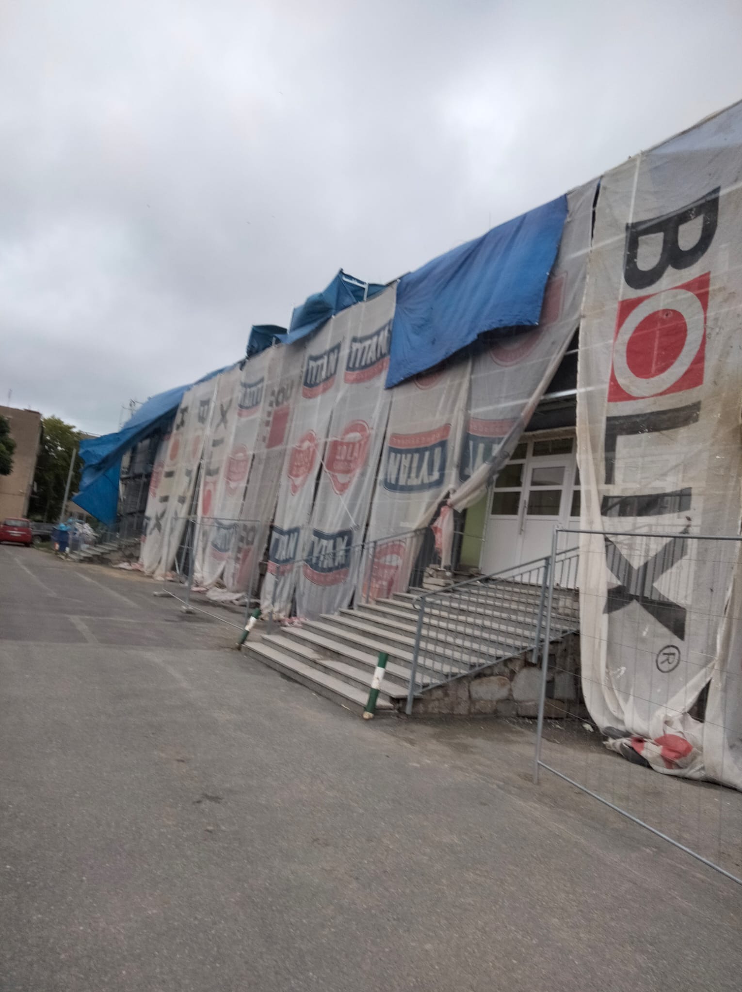 Budynek w trakcie renowacji, osłonięty reklamowymi plandekami, z widocznymi schodami wejściowymi i elementami rusztowania. Pochmurne niebo w tle.