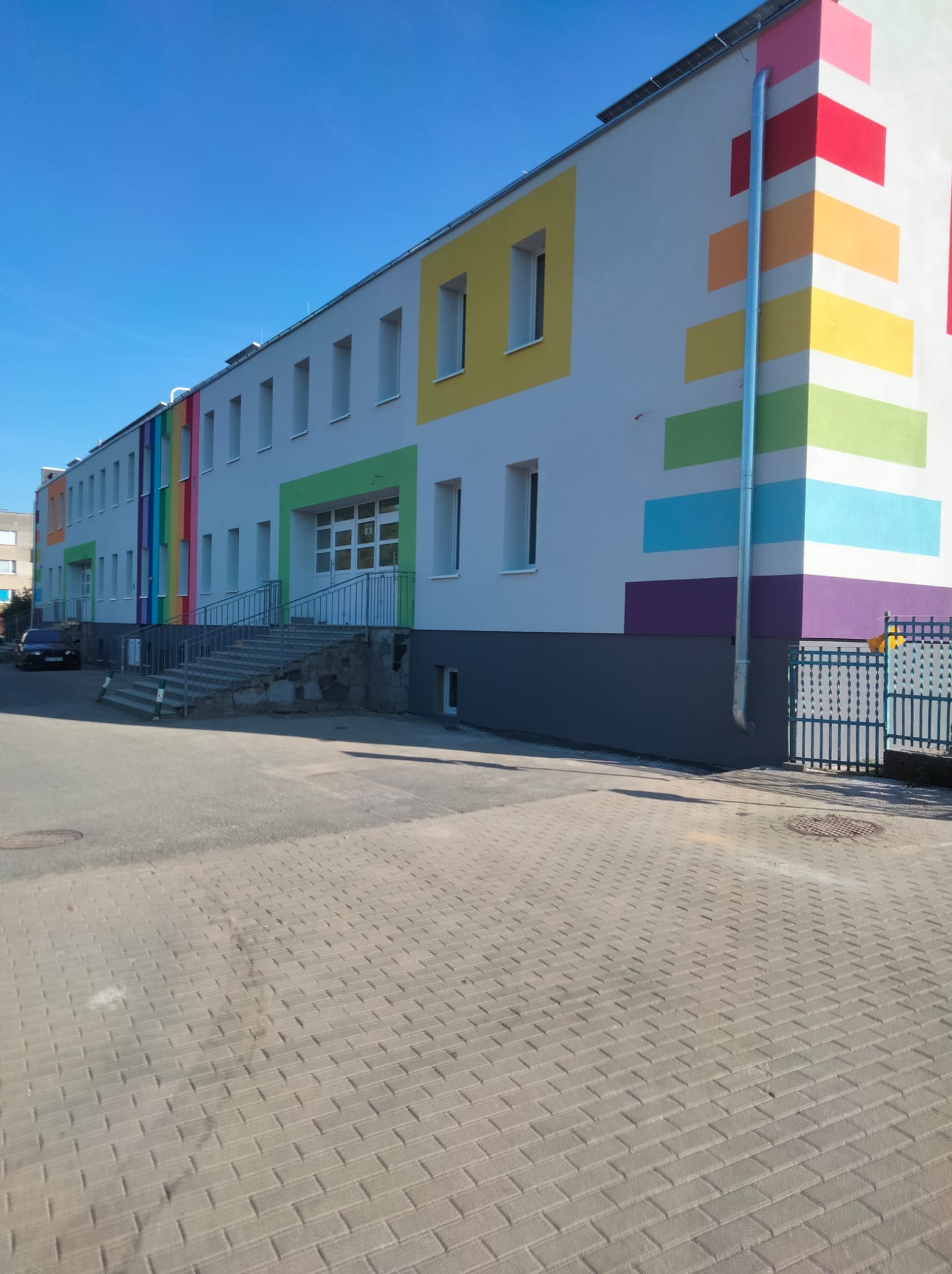 Kolorowa elewacja budynku z oknami i schodami wejściowymi. Fasada zdobiona poziomymi pasami w barwach tęczy, widoczne okna w kolorowych obramowaniach.