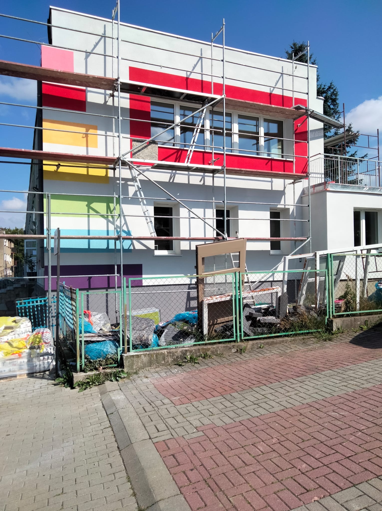 Dwupiętrowy budynek w trakcie renowacji elewacji z kolorowymi akcentami, otoczony rusztowaniami i ogrodzeniem z siatki. Widoczne okna i pozostałości po starych oknach.