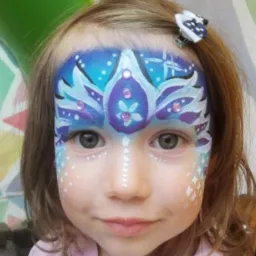 Dziewczynka z pomalowaną twarzą w niebiesko-fioletowe wzory z brokatem, przypominające koronę, z białą linią kropek na nosie i białymi wzorami na policzkach, w tle kolorowe dekoracje.